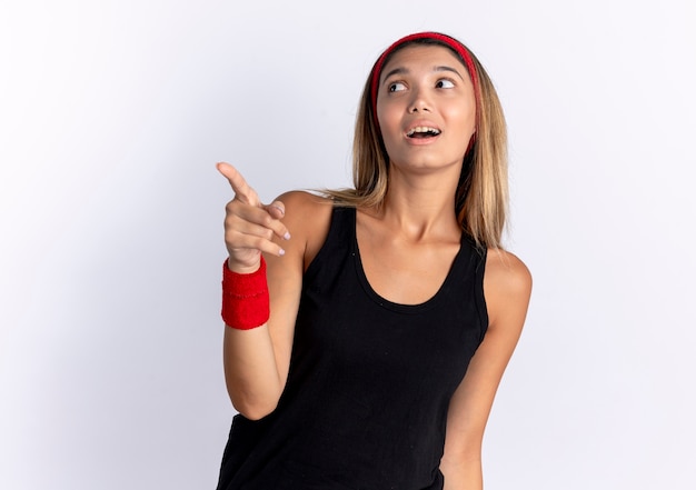 Young fitness girl in black sportswear and red headband looking confused pointign with finger at something scared standing over white wall