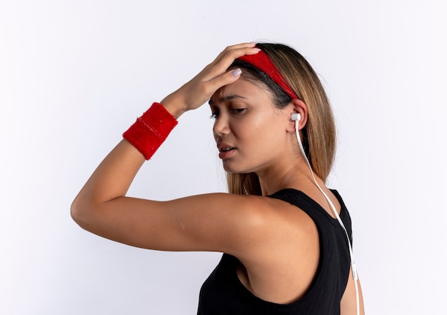 Young fitness girl in black sportswear and red headband looking confused with hand on her head for mistake standing over white wall