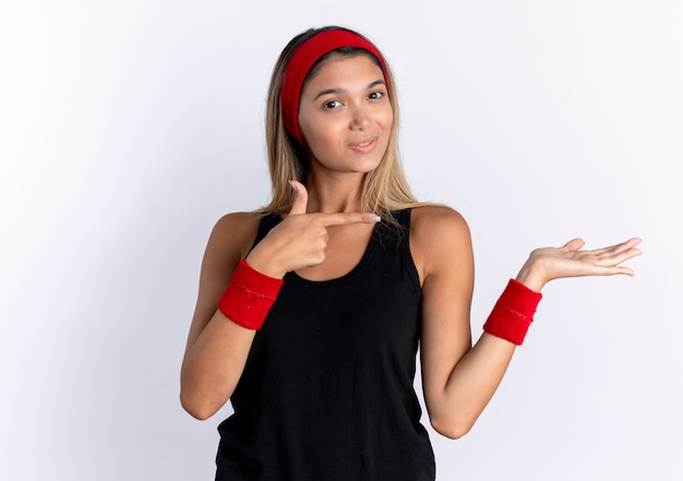 白い壁の上に立っている手の腕で何かを提示する側に指で指している黒いスポーツウェアと赤いヘッドバンドの若いフィットネスの女の子