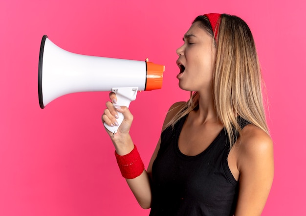 Young fitness girl in black sportswear and red headband shouting to megaphone over pink