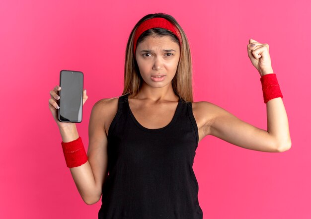 ピンクの壁の上に立っている怒っている顔で拳を握り締めるスマートフォンを示す黒いスポーツウェアと赤いヘッドバンドの若いフィットネスの女の子