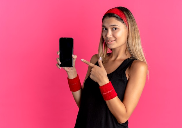 검은 운동복과 빨간 머리띠에 젊은 피트니스 소녀가 분홍색 위에 미소 짓는 손가락으로 스마트 폰 pointign을 보여주는