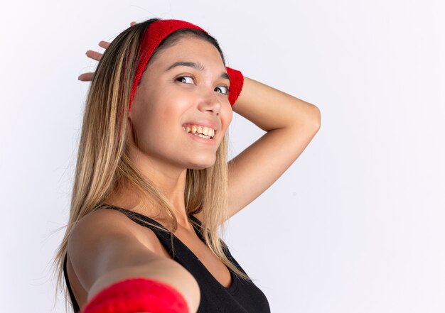 Young fitness girl in black sportswear and red headband smiling cheerfully standing over white wall