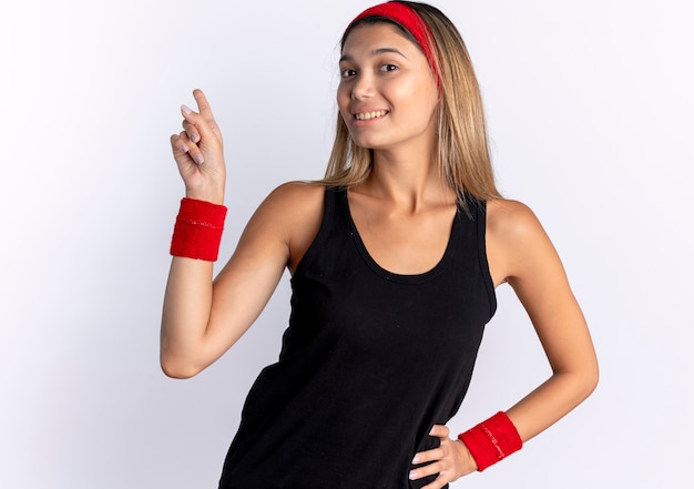 검은 운동복과 빨간 머리띠에 젊은 피트니스 소녀 흰색 벽 위에 서 검지 손가락으로 가리키는 자신감 미소