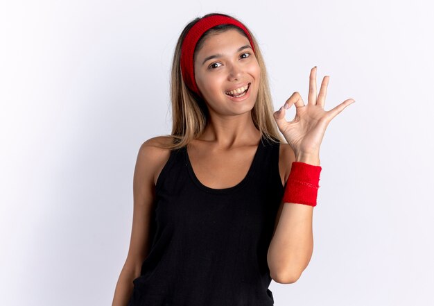 黒のスポーツウェアと赤いヘッドバンドの笑顔で白い壁の上に立っているOKのサインを示す若いフィットネスの女の子