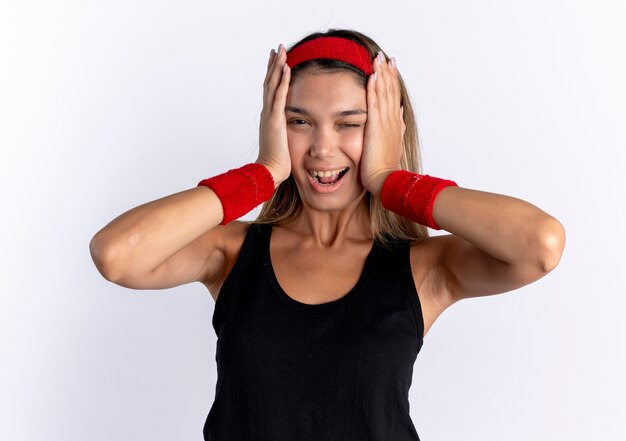 Молодая фитнес-девушка в черной спортивной одежде и красной повязке на голову удивлена и счастлива, стоя у белой стены
