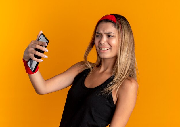 オレンジ色の壁の上に立って不機嫌そうに見えるスマートフォンを使用してselfieを取っている黒いスポーツウェアと赤いヘッドバンドの若いフィットネスの女の子