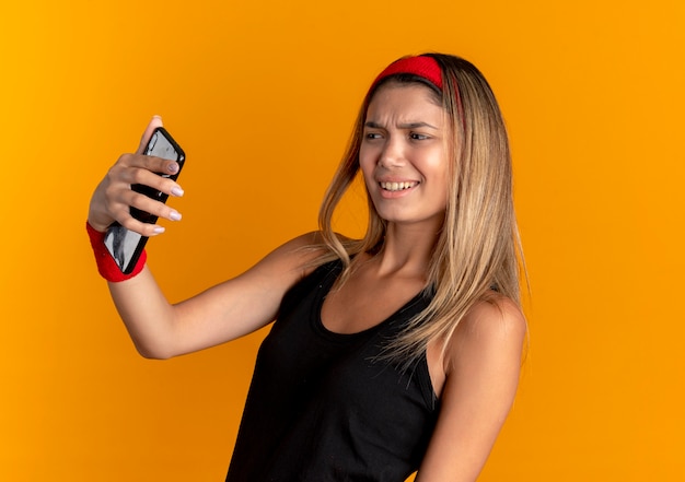 オレンジ色の壁の上に立って不機嫌そうに見えるスマートフォンを使用してselfieを取っている黒いスポーツウェアと赤いヘッドバンドの若いフィットネスの女の子