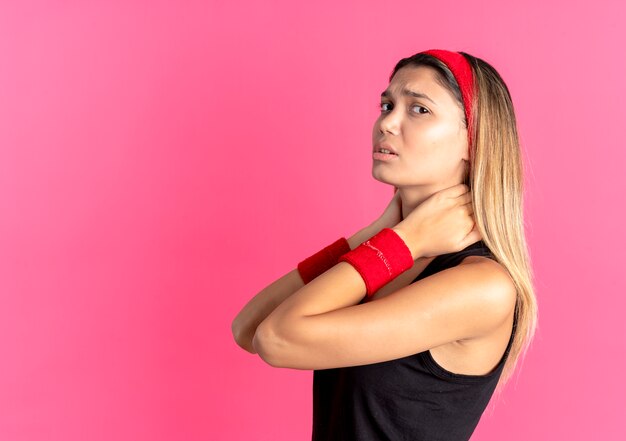 Молодая фитнес-девушка в черной спортивной одежде и красной повязке на голове, касаясь ее шеи, выглядит нездоровой, чувствуя боль над розовым