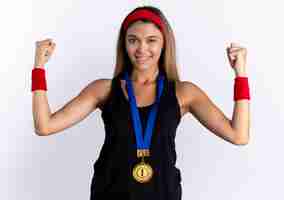 Free photo young fitness girl in black sportswear and red headband with gold medal around neck raising fists happy and positive winking and smiling standing over white wall