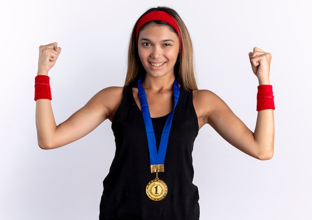 Young fitness girl in black sportswear and red headband with gold medal around neck raising fists happy and positive winking and smiling standing over white wall