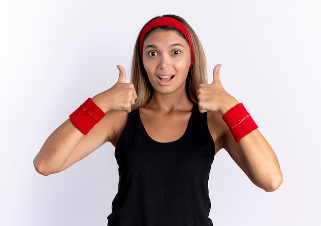 Молодая фитнес-девушка в черной спортивной одежде и красной повязке со счастливым лицом показывает палец вверх, стоя над белой стеной