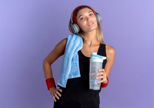 Young fitness girl in black sportswear and red headband with headphones and towel on shoulder holding bottle of water smiling confident over blue