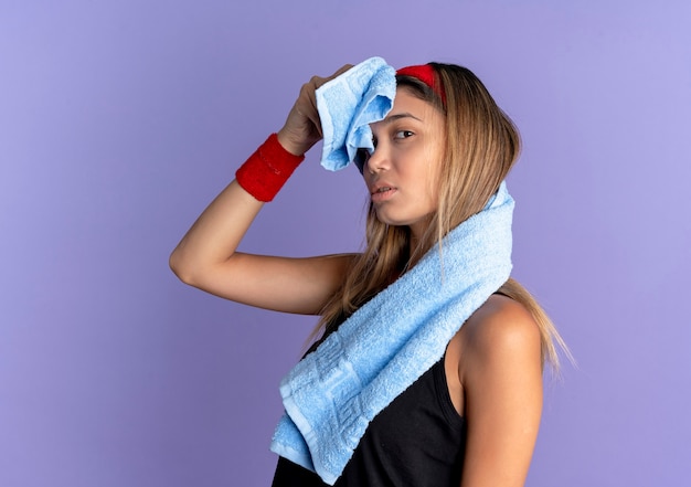 Foto gratuita giovane ragazza di forma fisica in abbigliamento sportivo nero e fascia rossa con le cuffie che pulisce la fronte con l'asciugamano che sta sopra la parete blu