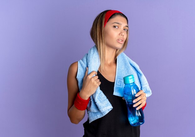 Young fitness girl in black sportswear and red headband with towel around neck  confused holding bottle of water standing over blue wall