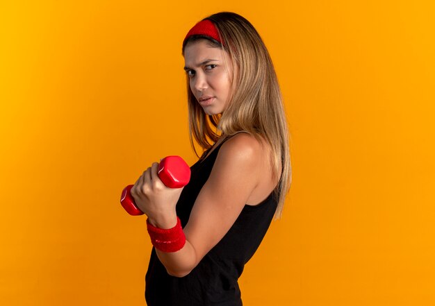 Молодая фитнес-девушка в черной спортивной одежде и красной повязке на голове, тренирующаяся с гантелями, уверенно выглядит над оранжевым