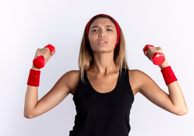 Young fitness girl in black sportswear and red headband working out with dumbbells looking confident standing over white wall