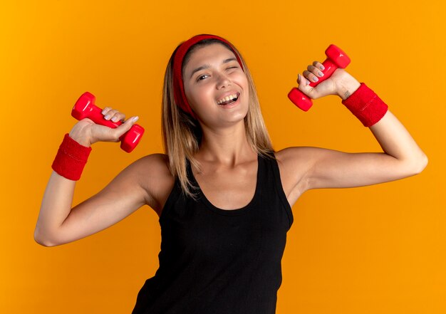 Young fitness girl in black sportswear and red headband working out with dumbbells smiling cheerfully  standing over orange wall