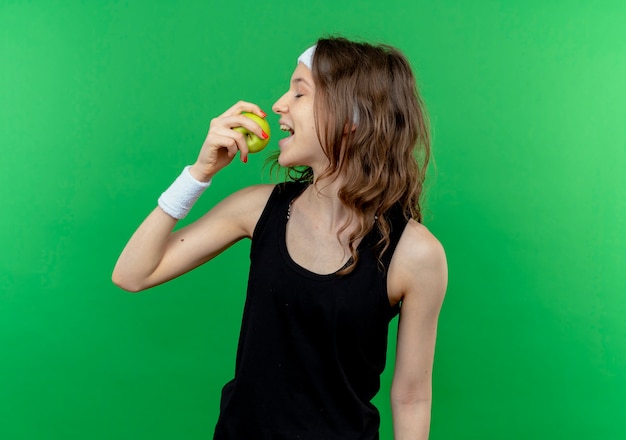 Foto gratuita giovane ragazza di forma fisica in abiti sportivi neri con fascia che morde la mela verde in piedi sopra la parete verde