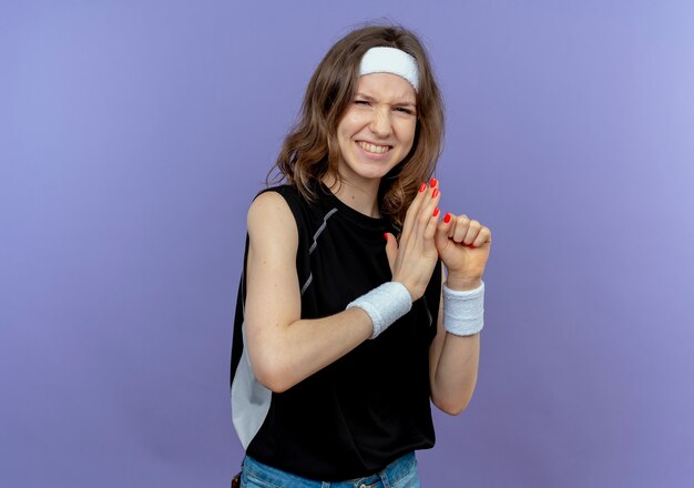 Молодая фитнес-девушка в черной спортивной одежде с оголовьем, сжимая кулак, очень эмоционально стоит над синей стеной