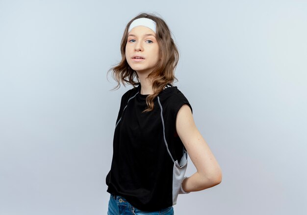 Young fitness girl in black sportswear with headband  confused standing over white wall