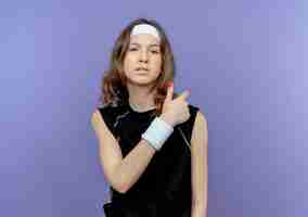 Free photo young fitness girl in black sportswear with headband displeased pointing back standing over blue wall