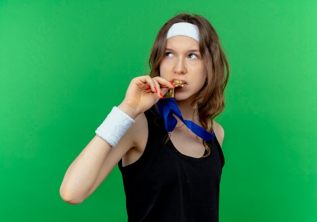 Young fitness girl in black sportswear with headband and gold medal around neck looking aside biting her medal standing over green wall