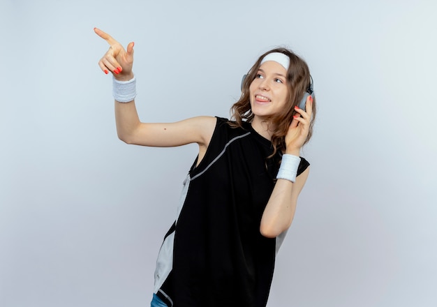 Young fitness girl in black sportswear with headband and headphones looking aside smiling pointing with finger at something standing over white wall