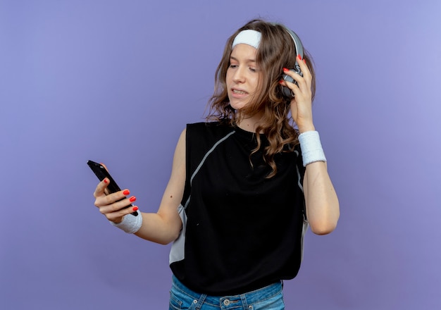 Молодая фитнес-девушка в черной спортивной одежде с повязкой на голову и наушниками, глядя на свой смартфон, ищет музыку, стоящую над синей стеной
