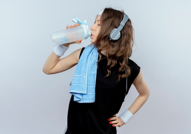 Free photo young fitness girl in black sportswear with headband and headphones and towel on shoulder drinking water after workout standing over white wall