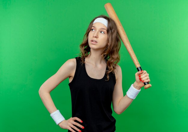 Young fitness girl in black sportswear with headband holding basaball bat hear head looking aside with pensive expression standing over green wall