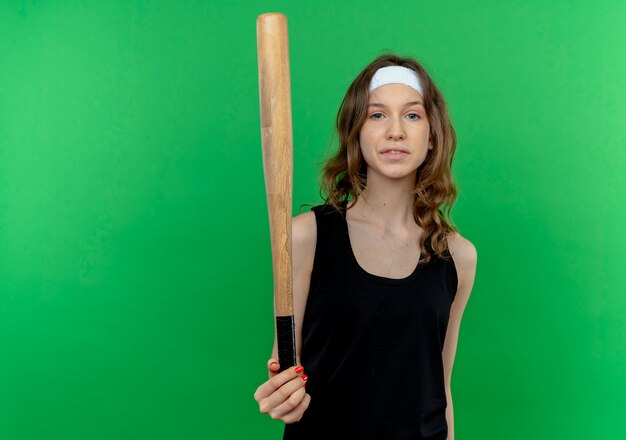 Young fitness girl in black sportswear with headband holding basaball bat  with confident expression standing over green wall