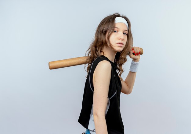 Молодая фитнес-девушка в черной спортивной одежде с повязкой на голову держит бейсбольную биту с серьезным лицом, стоящим над белой стеной