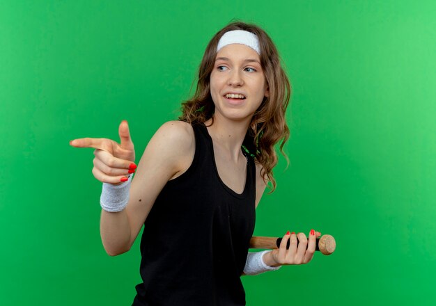 Young fitness girl in black sportswear with headband holding baseball bat pointing with index finger to the side standing over green wall