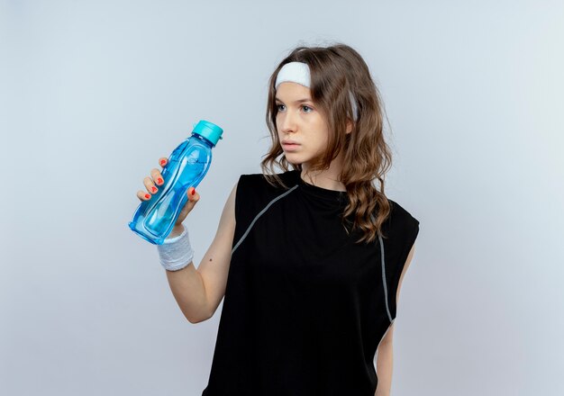 Молодая фитнес-девушка в черной спортивной одежде с повязкой на голову держит бутылку воды, озадаченно глядя в сторону, стоя над белой стеной