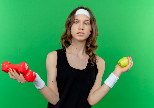 Young fitness girl in black sportswear with headband holding dumbbells and green apple looking confused standing over green wall