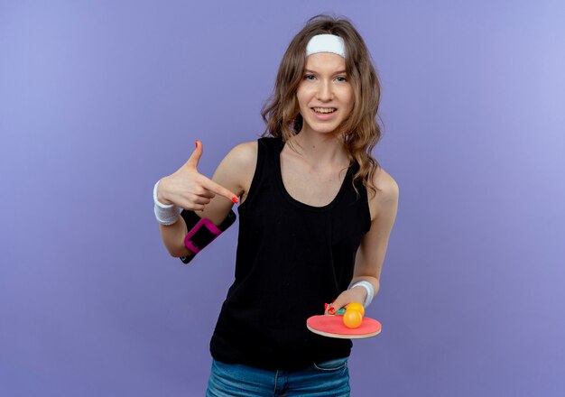 Young fitness girl in black sportswear with headband holding racket and balls for table tennis pointing with finger looking confident standing over blue wall