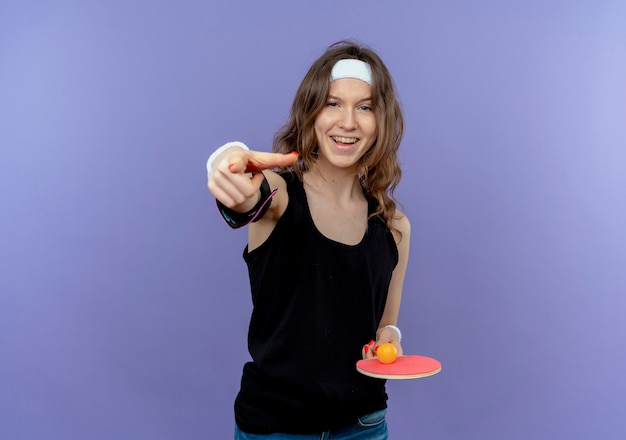 Young fitness girl in black sportswear with headband holding racket and balls for table tennis pointing with index finger smiling standing over blue wall
