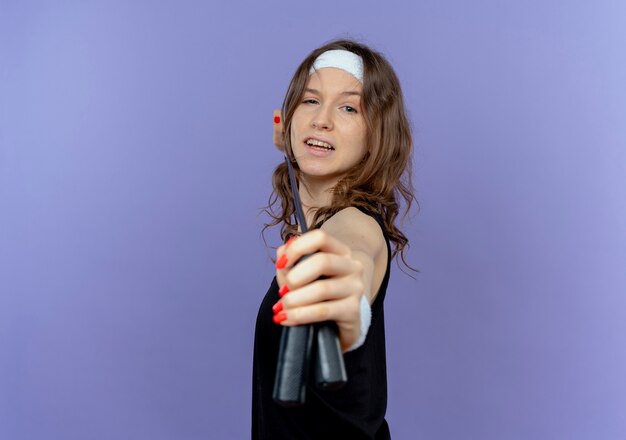 Young fitness girl in black sportswear with headband holding skipping rope like aiming with arrow and bow standing over blue wall