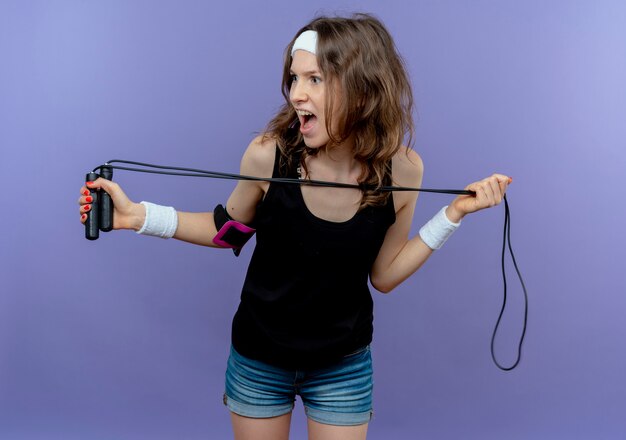 Young fitness girl in black sportswear with headband holding skipping rope looking aside scared standing over blue wall