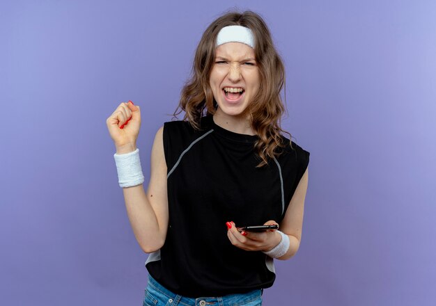 Young fitness girl in black sportswear with headband holding smartphone clenching fist happy and excited standing over blue wall