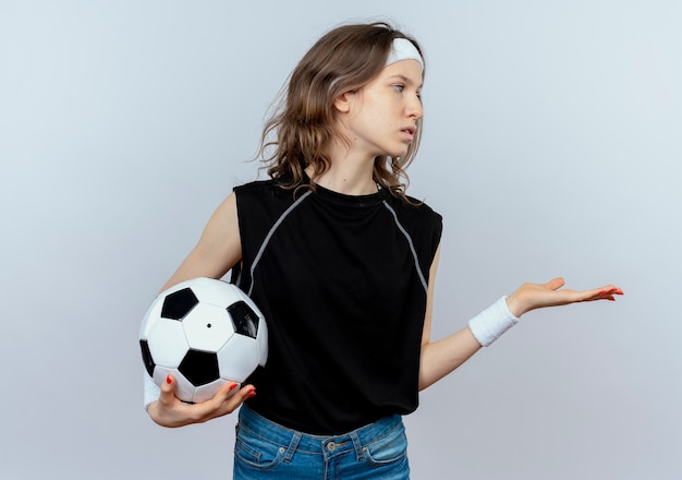 Young fitness girl in black sportswear with headband holding soccer ball looking aside with arm out as asking standing over white wall