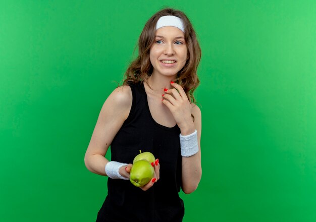 Young fitness girl in black sportswear with headband holding two green apples smiling cheerfully standing over green wall