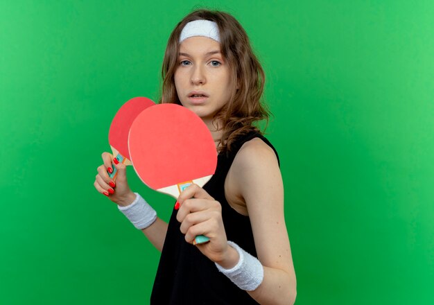 Young fitness girl in black sportswear with headband holding two rackets for table tennis  with serious face standing over green wall