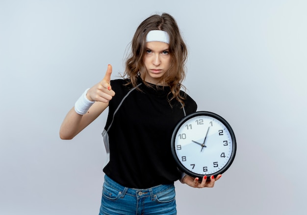 Young fitness girl in black sportswear with headband holding wall clock pointing with index finger with serious face standing over white wall