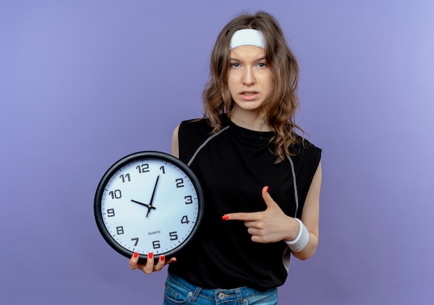 Free photo young fitness girl in black sportswear with headband holding wall clock pointing witn finger at it with serious face standing over blue wall