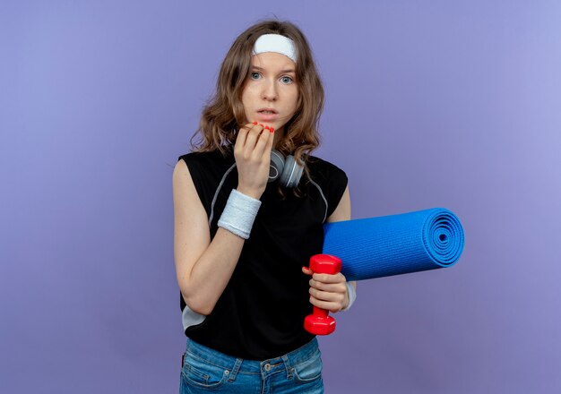 Young fitness girl in black sportswear with headband holding yoga mat and dumbbell worried and confused over blue