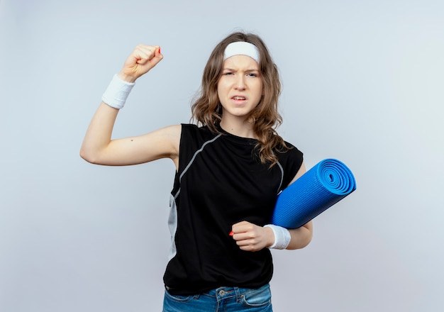 Молодая фитнес-девушка в черной спортивной одежде с повязкой на голову, держащая коврик для йоги, поднимая кулак, эмоциональное положение над белой стеной