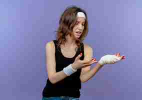 Free photo young fitness girl in black sportswear with headband lookign at her bandaged wrist being confuse and displeased standing over blue wall