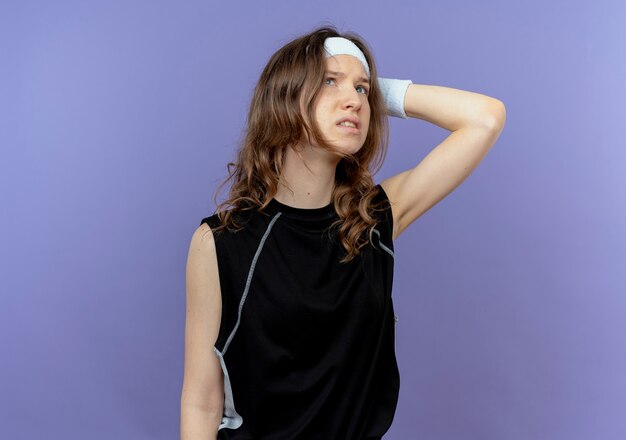 Young fitness girl in black sportswear with headband looking aside puzzled standing over blue wall